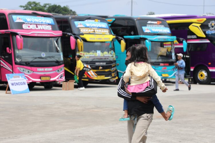 Seorang warga menggendong anaknya dengan latar belakang sejumlah bus siap mengantarkan untuk mudik. Dokumentasi Kemenhub.